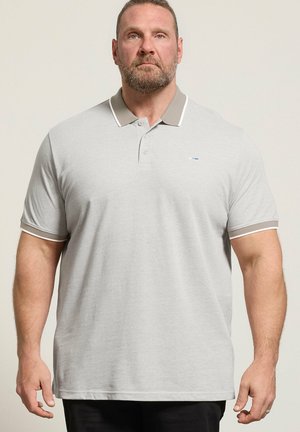 Dorosły mężczyzna z krótkimi włosami i brodą, ubrany w jasnoszary polo z kontrastowym kołnierzykiem i lamówką na rękawach, stojący na jednolitym tle.