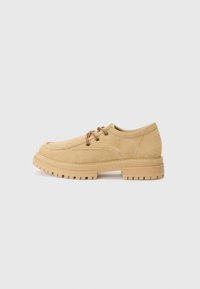 Suede lace-up shoes in a light tan color with a rounded toe, thick textured sole, and stitched detailing. Features brown laces and logo accents.