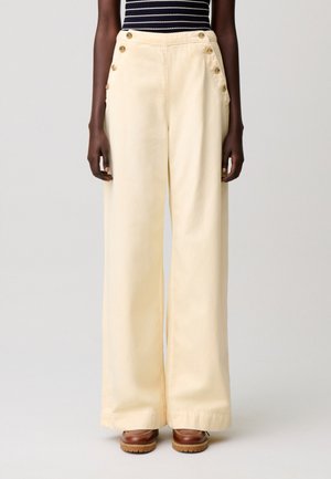 Mujer con pantalones anchos de talle alto en color crema con botones dorados en las caderas, combinados con zapatos de plataforma marrones y una camiseta de rayas negras y blancas.