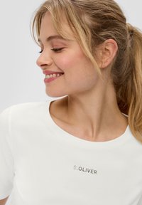 Weißes kurzärmliges T-Shirt mit rundem Halsausschnitt; auf der Vorderseite mit metallic „S. OLIVER“ Aufdruck. Glattes Baumwollmaterial.
