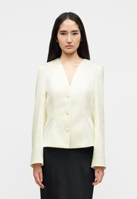 Frau mit langem dunklem Haar, die ein cremefarbenes, geknöpftes Blazer und einen schwarzen Rock trägt, stehend vor einem schlichten hellen Hintergrund.