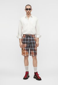 Homme portant des lunettes de soleil noires, une chemise blanche à manches longues, un short à carreaux orange et bleu usé, des chaussettes blanches et des bottes rouges à lacets, debout.