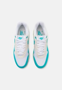 Ténis Nike Air com um upper em malha branca, detalhes em turquesa, atacadores redondos e sola em borracha texturizada. Design plano, silhueta de corte baixo.