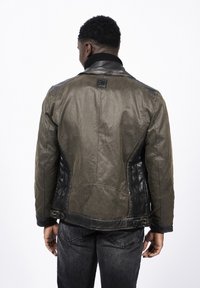 Mężczyzna zwrócony tyłem, ubrany w dwukolorową brązowo-czarną skórzaną kurtkę z klamrami oraz czarne jeansy na białym tle.