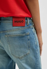 Jeans in denim azzurro con un'etichetta logo rossa con la scritta "HUGO" sul retro. Tra le caratteristiche ci sono tasche posteriori e cuciture beige.