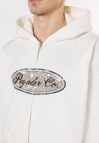 White zip-up hoodie featuring a large oval logo with "Pegador Co." in black and a textured brown background, soft material, and a spacious hood.
