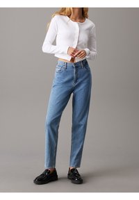 Weißer, gerippter, langärmliger Cropped-Cardigan mit Knopfverschluss, kombiniert mit hellblauen, lässig geschnittenen Jeans und schwarzen Schuhen.