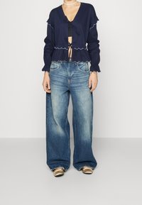 Giacca cropped blu navy con bordi arruffati e chiusura a cravatta, abbinata a jeans in denim light blue a gamba larga. Design semplice, tessuto leggero.