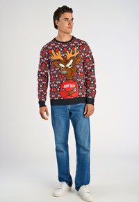 Czerwony sweter z dzianiny z festiwalowym motywem renifera, wzornikowany w płatki śniegu, z czarną talią i mankietami, noszony z niebieskimi dżinsami.