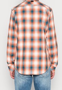 Chemise à manches longues à carreaux orange, bleu sarcelle et blanc. Dotée d'un col boutonné et d'un ourlet arrondi. Tissu léger et lisse avec un motif à carreaux.