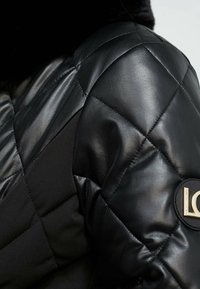 Svart vadderad jacka som består av läder och tyg, med en rund logotypdetalj på axeln och kontrasterande matt och blank yta.