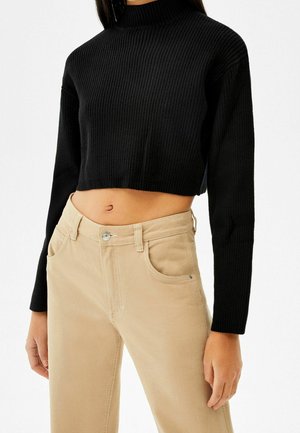 Pull en maille côtelée noir, court et à col haut. Associé à un pantalon beige avec poches avant et fermeture à bouton.