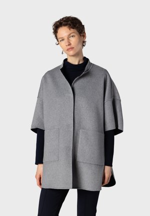 Femme portant un manteau ample gris à manches courtes sur un haut noir à manches longues et un pantalon noir, debout devant un fond clair uni.