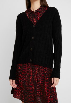 Femme portant un cardigan noir boutonné sur une robe rouge et noire à manches longues à motifs, debout devant un fond uni.