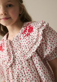 Weißes Kleid mit roten Blumenmuster und bestickten Akzenten, ausgestattet mit einem Rüschencollier und kurzen puffierten Ärmeln. Durchgehende Knopfverschluss vorne.