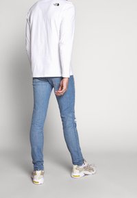 Bílá košile s dlouhými rukávy má malé logo, které je kombinováno se světle modrými skinny džíny a bílými teniskami s šedými a žlutými akcenty.