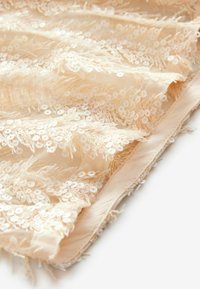 Tissu en dentelle beige avec des sequins blancs éparpillés et une broderie florale. Le bord présente des fils bruts, créant une finition texturée.
