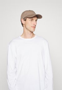 Beige Kappe mit einem gebogenen Schirm, getragen von einer Person in einem weißen Langarmhemd. Glattes Material, schlichtes Design, keine sichtbaren Muster oder Akzente.