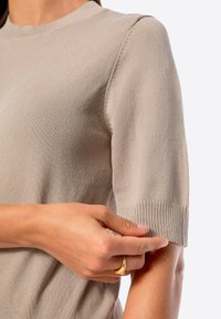 Beżowa, krótkosleeve swetrowa bluzka z okrągłym kołnierzykiem. Posiada prążkowane mankiety i gładką teksturę, podkreślając elastyczność i dopasowanie materiału.