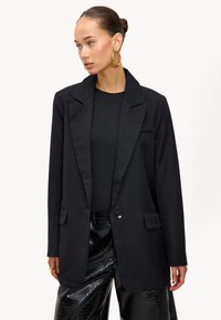 Zwarte blazer met een satijnen afwerking, met Engelse revers, twee voorzakken en een enkele knoopsluiting, gedragen over een zwarte top en nepleren broek.