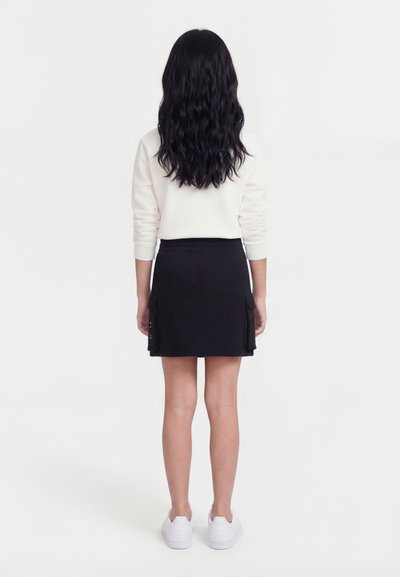 Κορίτσι με μακριά κυματιστά μαύρα μαλλιά στέκεται γυρισμένη προς τα πίσω, φορώντας λευκό μπλουζάκι με μακριά μανίκια, μαύρη φούστα με τσέπες και λευκά αθλητικά παπούτσια.