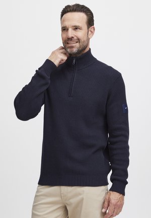 Marineblå ribbestrikket genser med glidelås i kragen, med et lite logo-lapp på venstre erme og teksturert, elastisk stoff.
