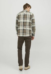 Camisa de franela a cuadros en tonos beige, negro y blanco. Presenta mangas largas, puños con botones y un dobladillo redondeado, combinada con pantalones marrones.