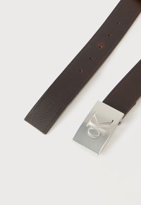 Σκούρο καφέ δερμάτινη ζώνη με ασημί ορθογώνιο αγκράφα χαραγμένη με τα γράμματα "CK" σε ανοιχτό φόντο.