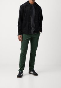 Man iklädd svart kordaroyajacka, mörk t-shirt, gröna kordaroybyxor och svarta sneakers med vita ränder, står på enfärgad vit bakgrund.