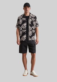 Schwarzes Kurzarmhemd mit weißem Blättermuster, kombiniert mit schwarzen Cargo-Shorts und weißen Sneakers. Enthält eine schwarze Kappe.