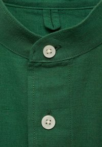 Green linen shirt with a Mandarin collar, featuring a white button and vertical stitching details on the placket. Texture appears soft and casual.