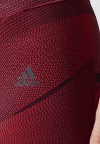 Czerwone legginsy sportowe w wzór, charakteryzujące się gładką teksturą, szarym logo Adidas oraz ciemnymi akcentami, w dopasowanym kroju.