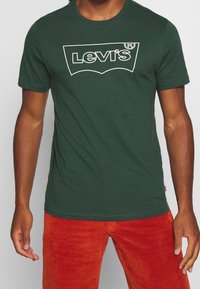 Zelené bavlněné tričko s bílým logem Levi's, krátkými rukávy a kulatým výstřihem, kombinované s oranžovými manšestrovými kalhotami.