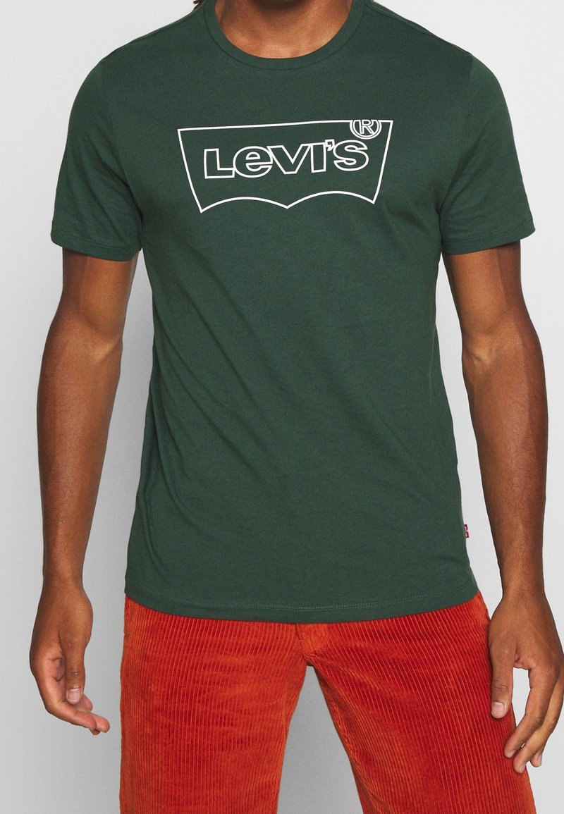 Zelené bavlněné tričko s bílým logem Levi's, krátkými rukávy a kulatým výstřihem, kombinované s oranžovými manšestrovými kalhotami.