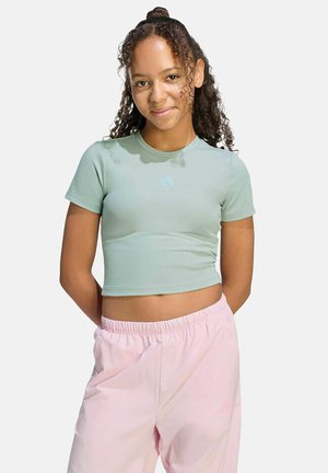 Adolescente aux cheveux bouclés portant un t-shirt court vert clair et un pantalon à taille élastique rose, souriante avec les mains derrière le dos.