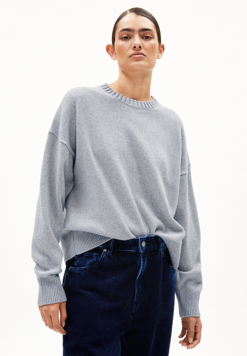 Frau in einem hellgrauen oversized Pullover und einer dunkelblauen Cordhose, die vor einem schlichten weißen Hintergrund steht.