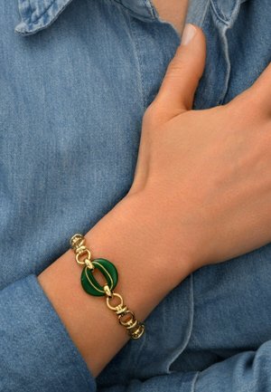 Bracelet doré avec des accents circulaires verts, design en chaîne liée et texture lisse. Porté sur une chemise en denim bleu clair.