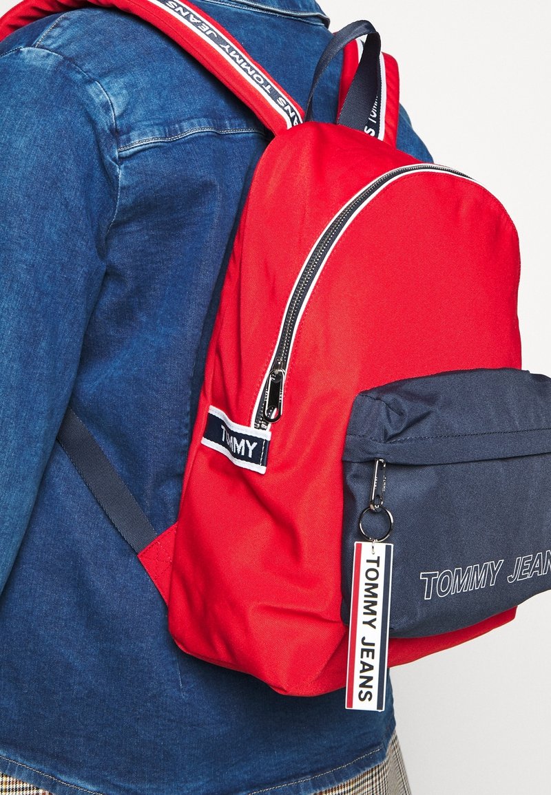 Personne portant une veste en jean bleu porte un sac à dos Tommy Jeans rouge et bleu marine avec des fermetures éclair de marque et un détail porte-clés.