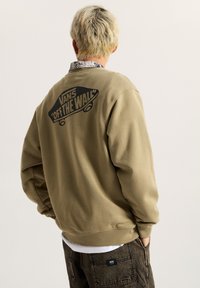Sudadera caqui con un logo negro "VANS OFF THE WALL" en la parte posterior. Presenta un corte relajado y puños acanalados, utilizada sobre una camiseta blanca.