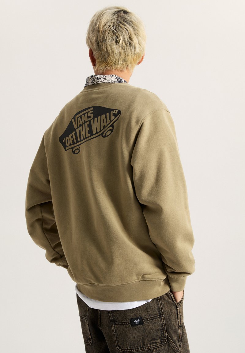 Sudadera caqui con un logo negro "VANS OFF THE WALL" en la parte posterior. Presenta un corte relajado y puños acanalados, utilizada sobre una camiseta blanca.