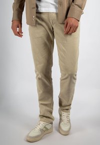 Man draagt beige corduroy broek, witte en grijze sneakers, een wit T-shirt en een deels dichtgeknoopte tan overhemd.