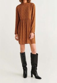 Braunes langärmeliges Kleid mit tailliertem Oberteil und ausgestelltem Rock, versehen mit einem schwarzen Punkte-Muster. Kombiniert mit knielangen schwarzen Stiefeln mit Absatz.