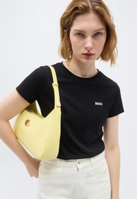 Νέα γυναίκα με κοντά ξανθά μαλλιά, φορώντας μαύρο t-shirt BOSS και λευκό παντελόνι, κρατώντας μια κίτρινη τσάντα ώμου σε απλό φόντο.