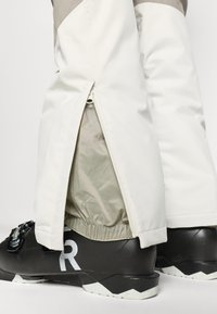 White waterproof ski pants with gray accents, featuring a zippered lower leg opening and elastic cuffs, paired with black ski boots.