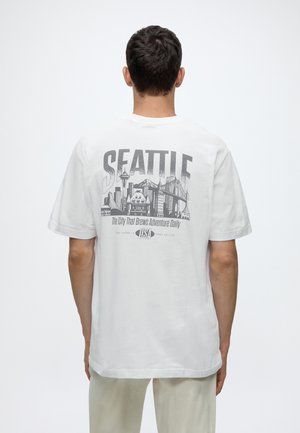 Mann von hinten gesehen, trägt ein weißes T-Shirt mit einer schwarzen Skyline von Seattle und Text über die Stadt auf dem Rücken.