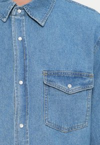 Jasnoniebieska koszula dżinsowa z ostrym kołnierzykiem, patką z guzikami z przodu i kieszenią na piersi. Charakteryzuje się fakturowaną tkaniną i zaokrągloną klapką kieszeni.