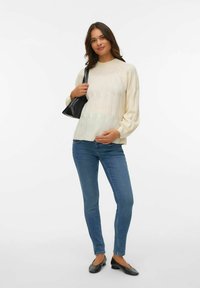 Kremasti pleteni pulover s rebrastom teksturom, napuhanim rukavima, uparen s plavim uskim trapericama i crnim balerinkama, drži malu crnu torbicu.