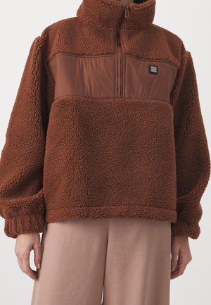 Brown sherpa fleece pullover with high collar, half-zip, and smooth fabric chest panel worn over beige pants.