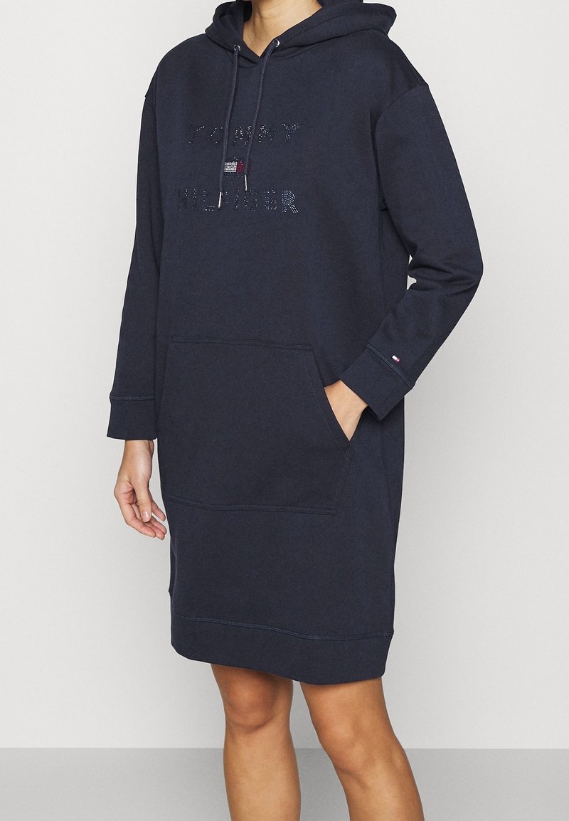Femme portant une robe à capuche Tommy Hilfiger bleu marine avec poche avant et capuche, main dans la poche, sur fond clair uni.
