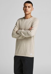 Beige long-sleeve knit shirt with a textured finish, crew neckline, and ribbed accents. Paired with dark trousers, model stands with arms crossed.
